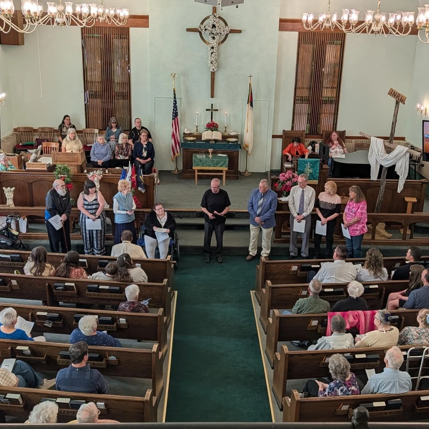 A church interior during a service or gathering. Several people are standing at the front, some holding papers, while others are seated or standing in the pews. The altar is decorated with flowers, flags, a cross, and an open Bible. The congregation is watching, and the room is lit by chandeliers.