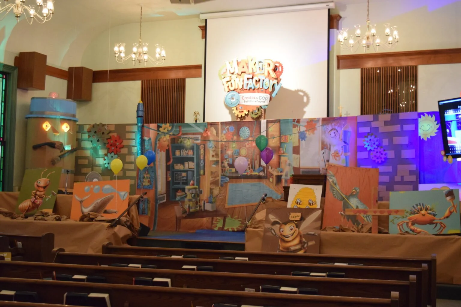 Decorated church interior with a stage set for a children's event, featuring cartoon bug characters, colorful balloons, artwork, and a sign that reads "Maker Fun Factory".
