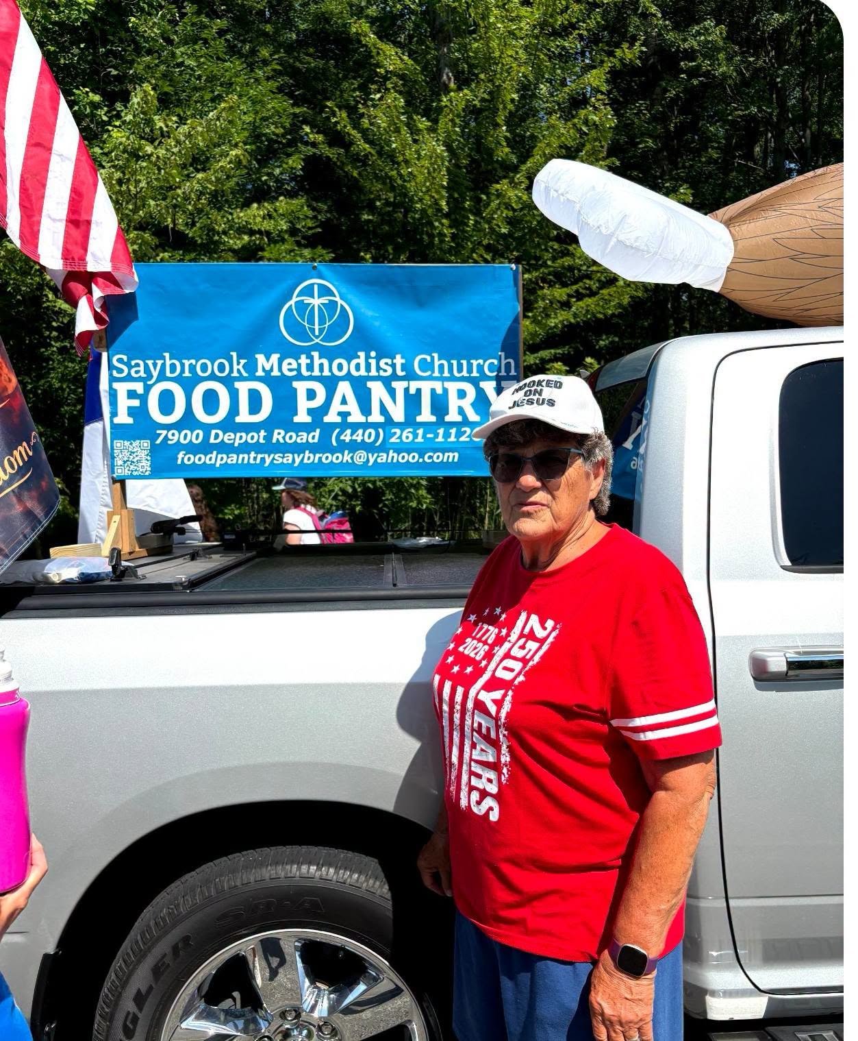 A woman wearing sunglasses, a white cap, and a red shirt standing next to a silver pickup truck with a Saybrook Methodist Church Food Pantry banner in the background. The banner provides the address and contact information for the food pantry. The woman is outdoors with green trees behind her.