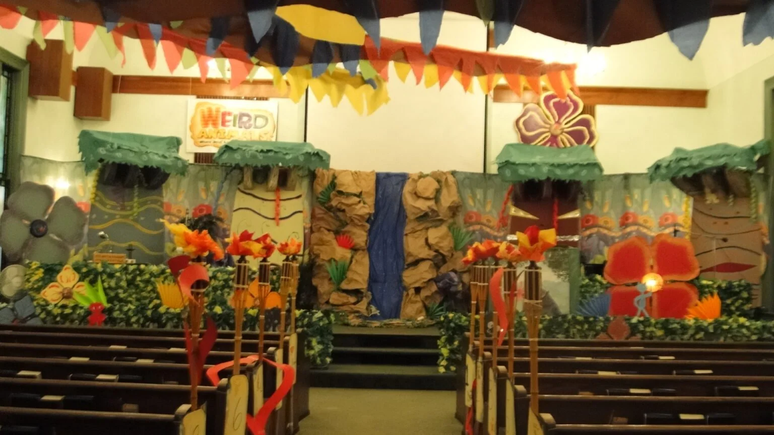 Decorated stage with tropical theme, featuring a waterfall, large flower cutouts, colorful banners, and foam flowers. The stage is flanked by rows of chairs and festive foliage.