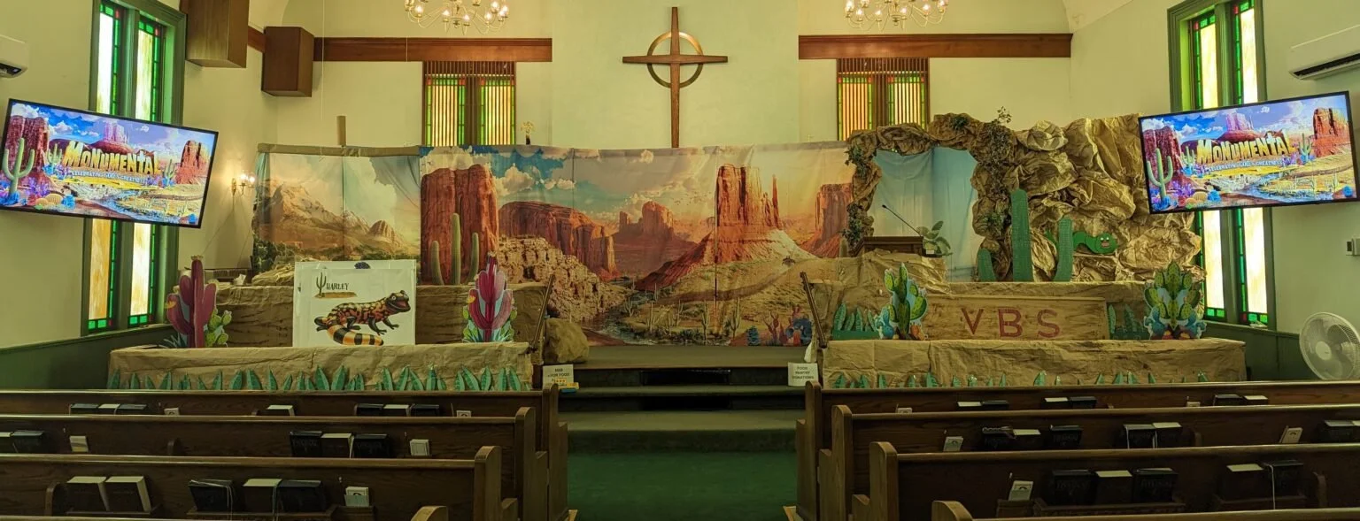 Church interior decorated with a desert scene backdrop, cactus cutouts, and a large cross on the wall. There are two TVs on either side displaying a colorful scene with the text 'El Monumetal'.