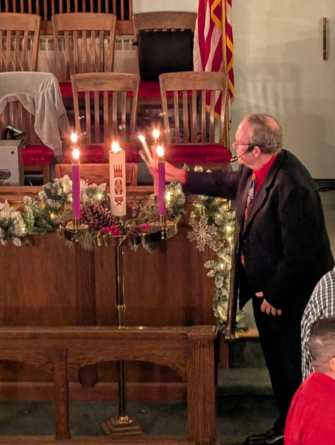 A man lighting candles on an Advent wreath decorated with pinecones, flowers, leaves, and lights, during a Christmas celebration. The wreath features four candles, three purple and one pink. The background includes wooden chairs, a table with a tablecloth, and a flag.