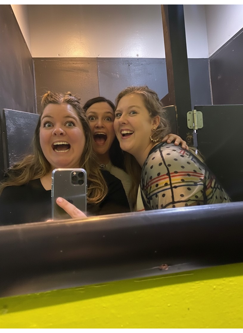Three women taking a selfie together in a mirror, showing happy expressions, inside a small, dimly lit room or bathroom. Besties. Grief. Kindred Spirits.