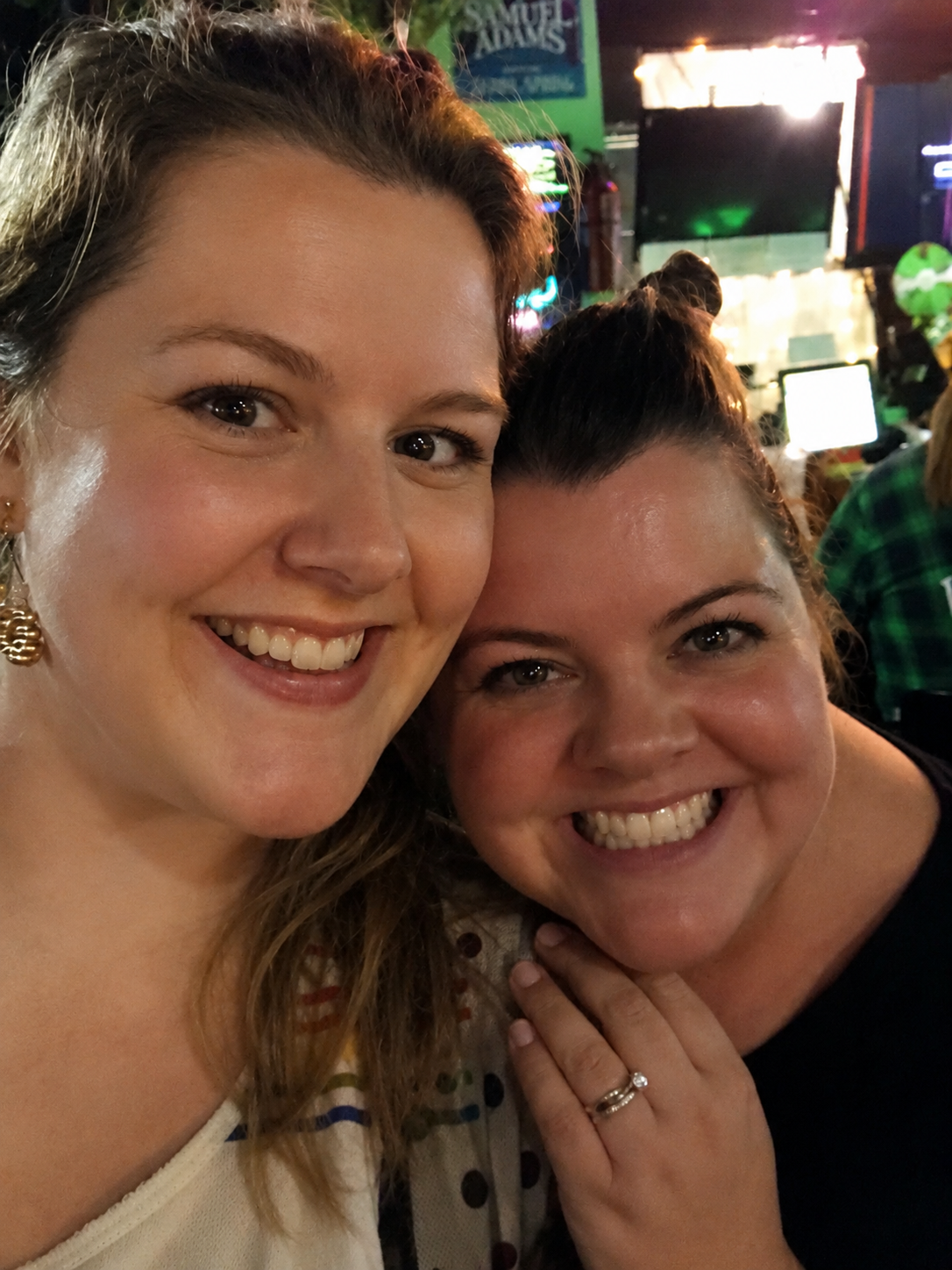 Two women smiling and posing for a selfie at a lively indoor event or bar, with colorful lights and screens in the background.