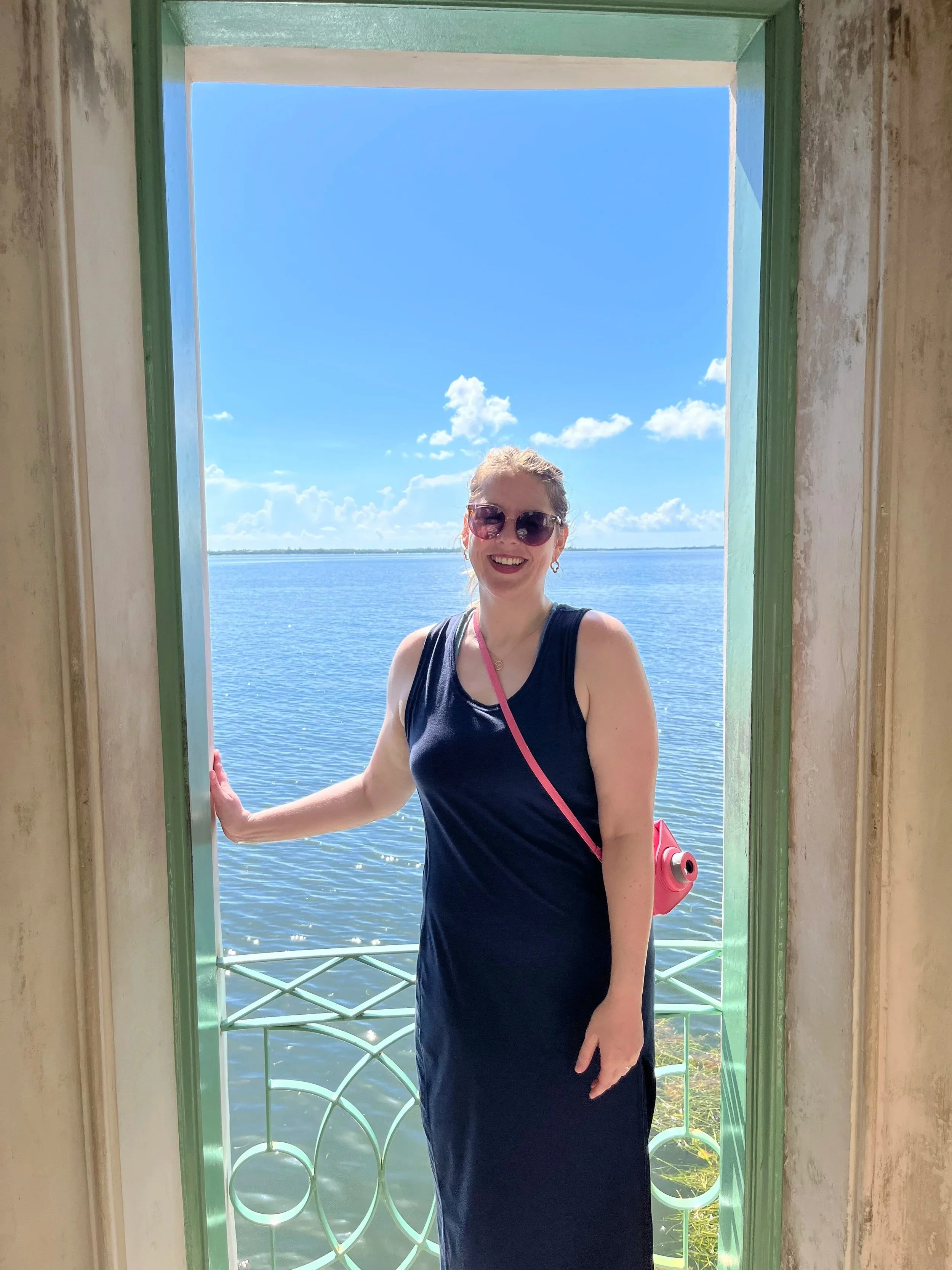 A woman with blonde hair, sunglasses, and earrings, wearing a black dress and pink bag, standing at a window opening with a view of a blue lake and partly cloudy sky. Polaroid camera, Vizcaya Museum and Gardens in Miami, FL.