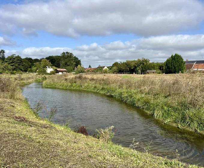 Rivière calme traversant un paysage rural avec des champs verts et des maisons en arrière-plan, sous un ciel partiellement nuageux.