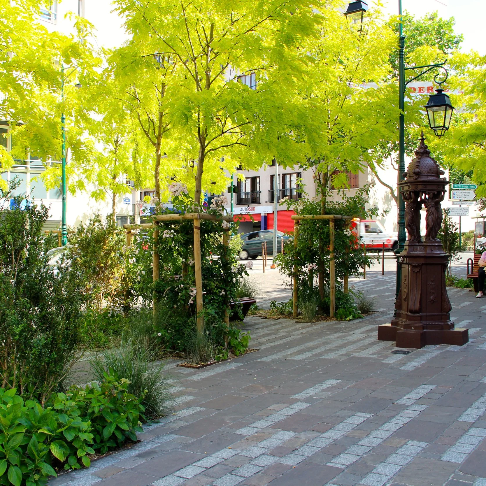 Un parc urbain avec des arbres feuillus verts, une fontaine en métal ornée de figures humaines, un lampadaire ancien,  un trottoir pavé, des immeubles résidentiels en arrière-plan et des voitures en mouvement.
