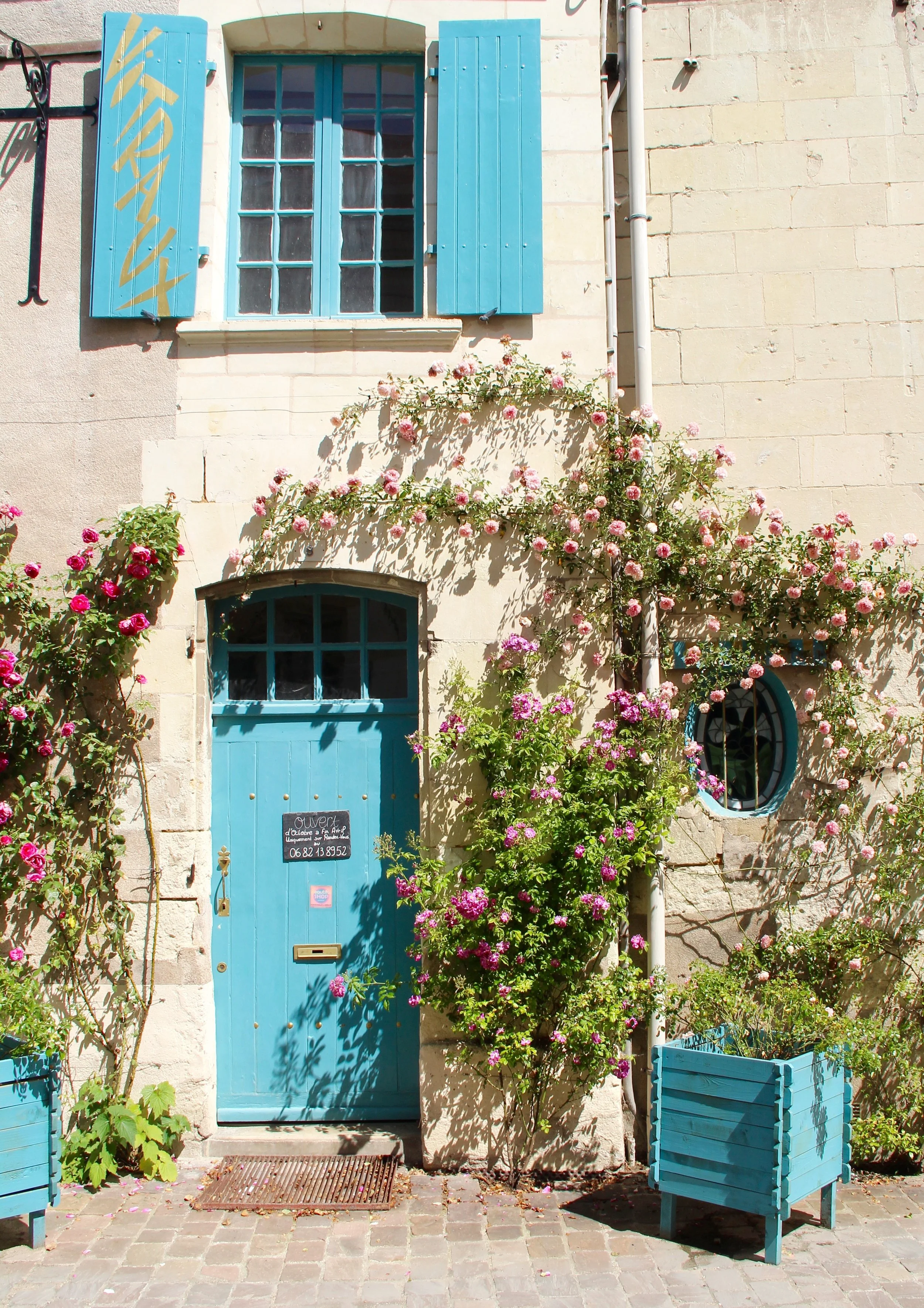 Une façade de maison en pierre avec une porte bleue et des volets de fenêtre bleus. Il y a des plantes et des fleurs roses et violettes autour de la porte, avec deux grands contenants en bois peint en bleu également.