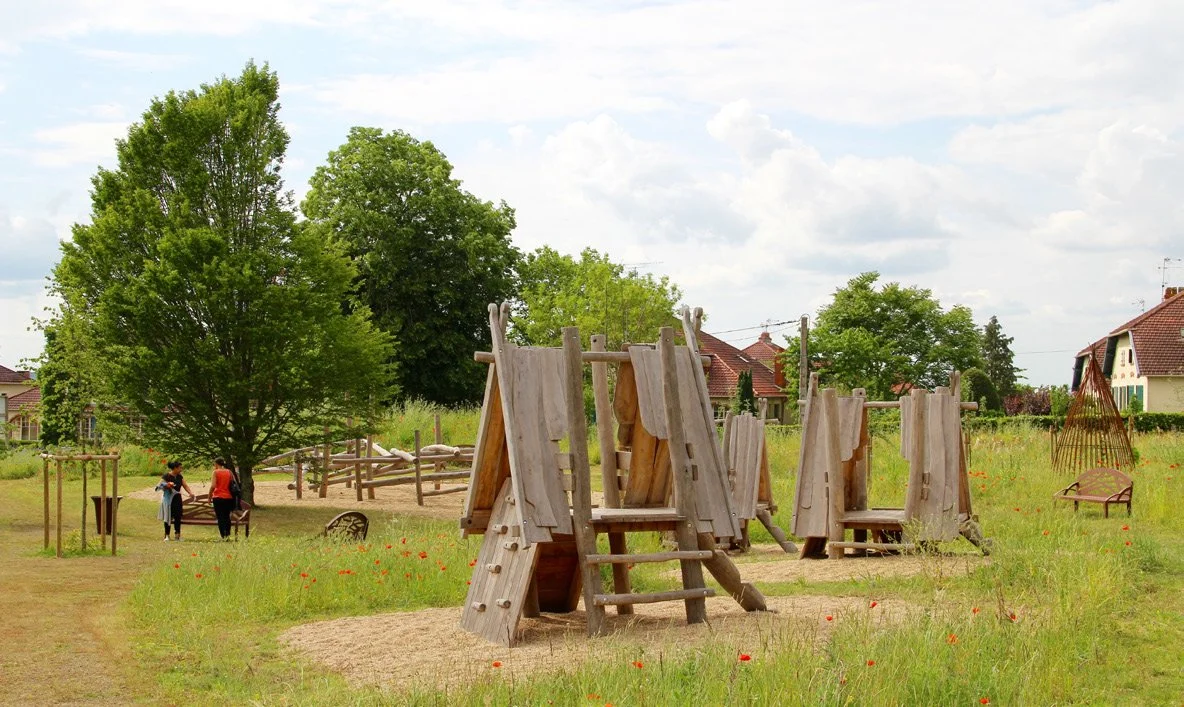 A playground with wooden structures in a grassy area with trees and houses in the background, and children playing.