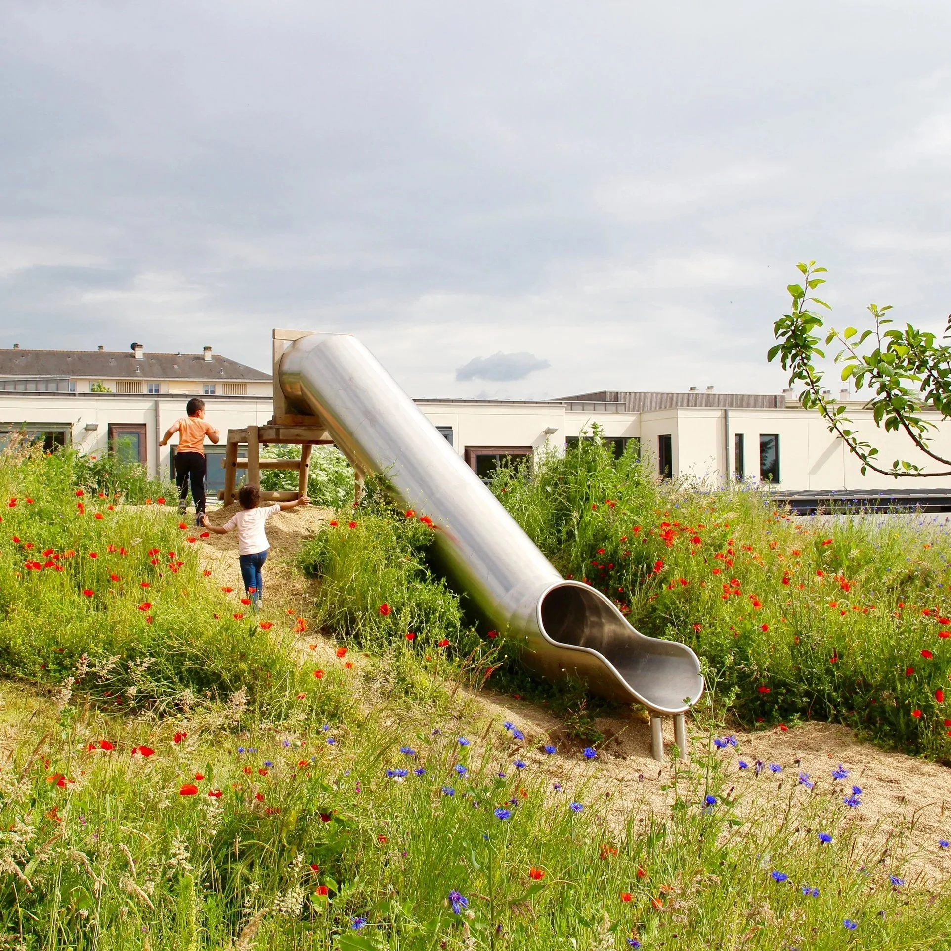 Enfant jouant dans un jardin avec une grande glissière en métal, entouré de fleurs colorées et d'arbres, avec des bâtiments modernes en arrière-plan et un ciel nuageux.