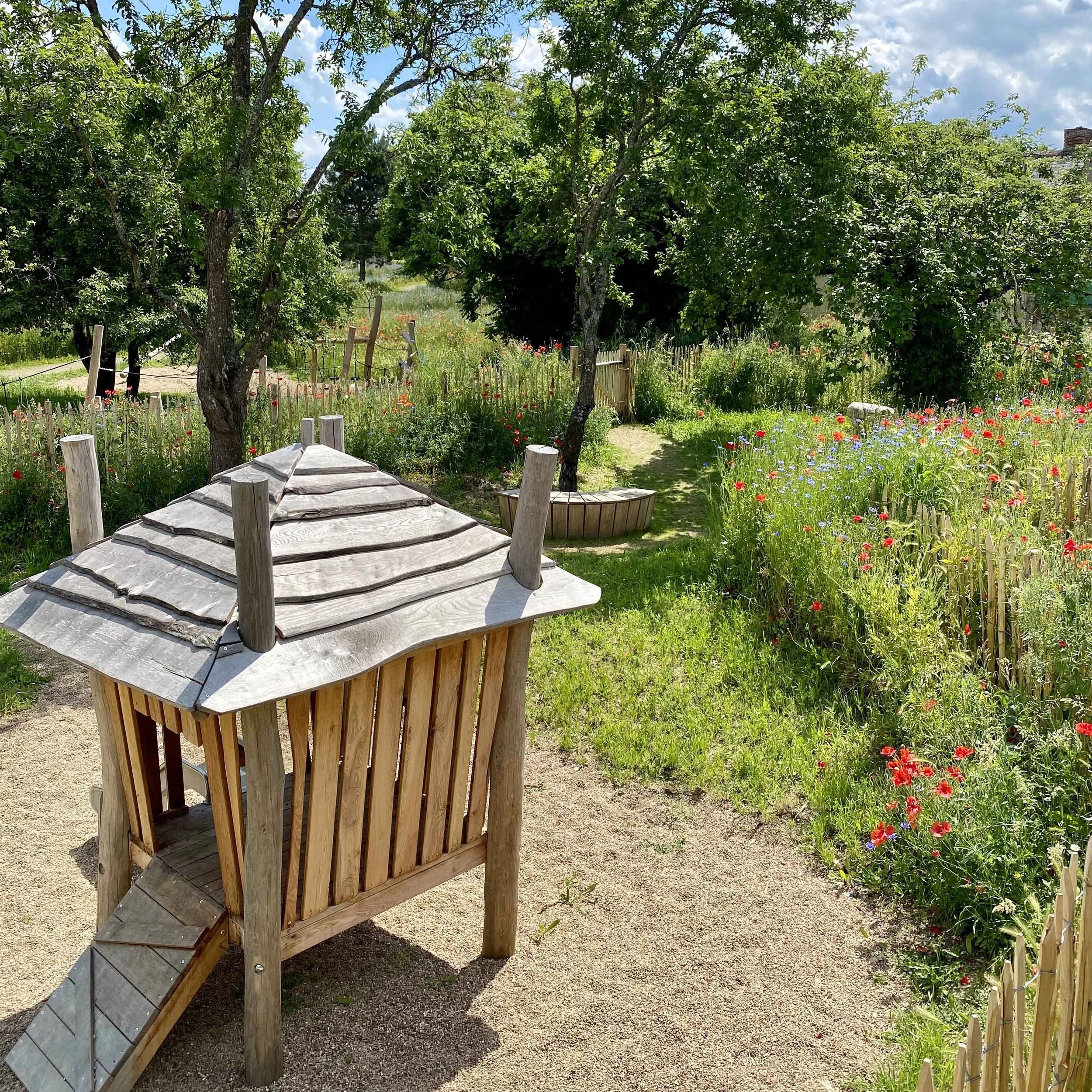 Une cabane en bois dans un jardin fleuri avec des arbres et un ciel partiellement nuageux.