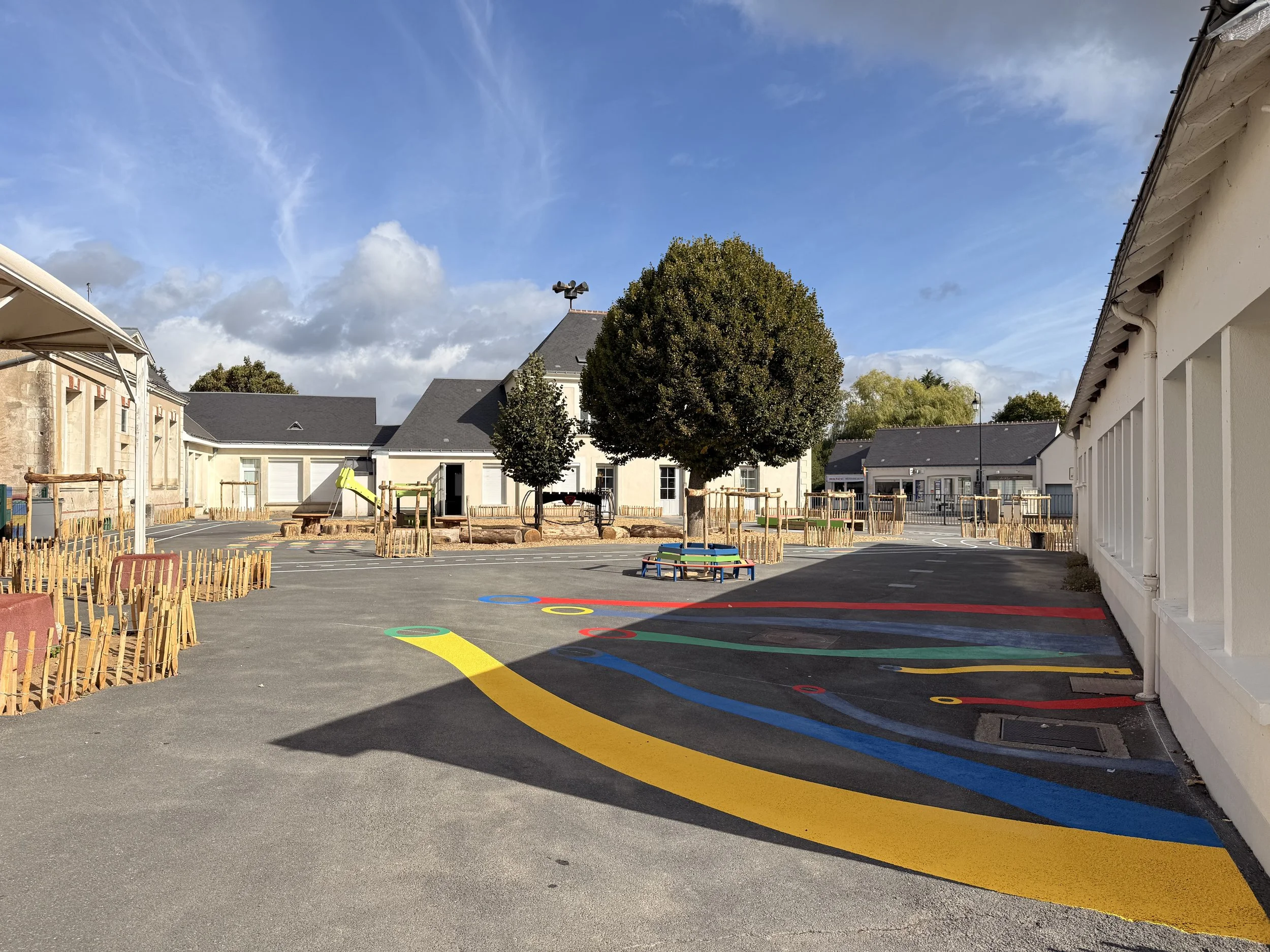 Une cour d'école avec des peintures colorées au sol et des aires de jeu, entourée de bâtiments à un ciel bleu avec quelques nuages.