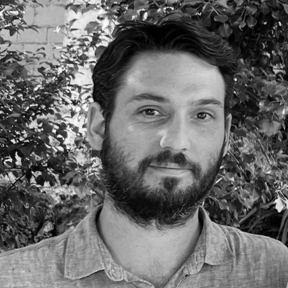 Portrait d'un homme avec une barbe, portant une chemise, se tenant devant des plantes et des feuilles.