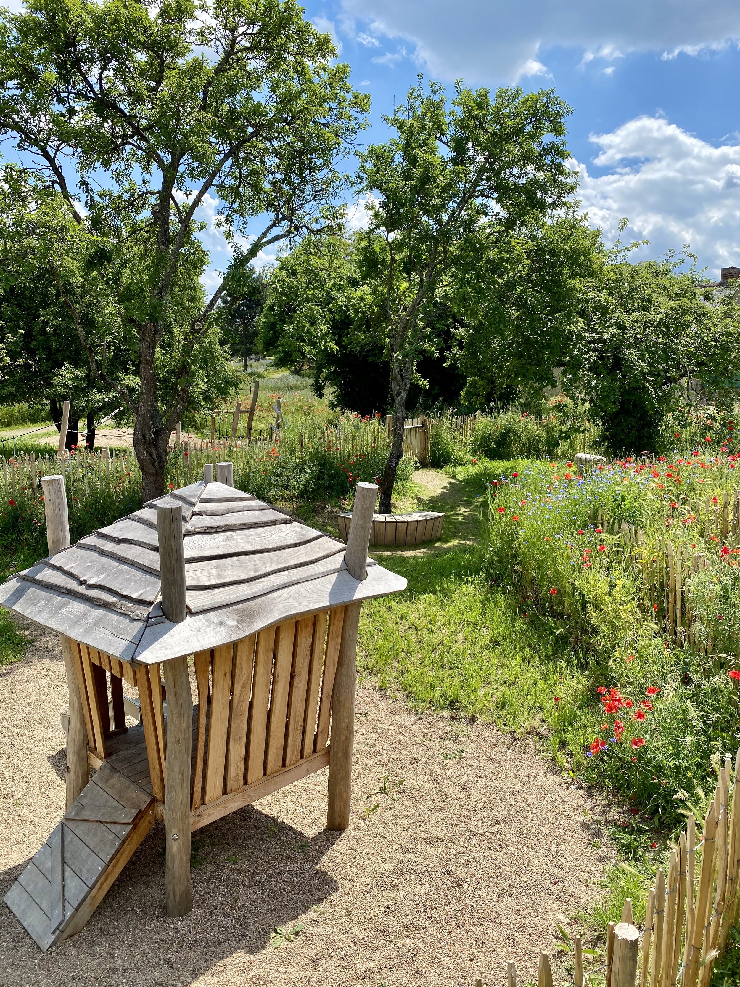 Jardin avec des arbres, des fleurs colorées et une petite clôture en bois, sous un ciel bleu avec des nuages.