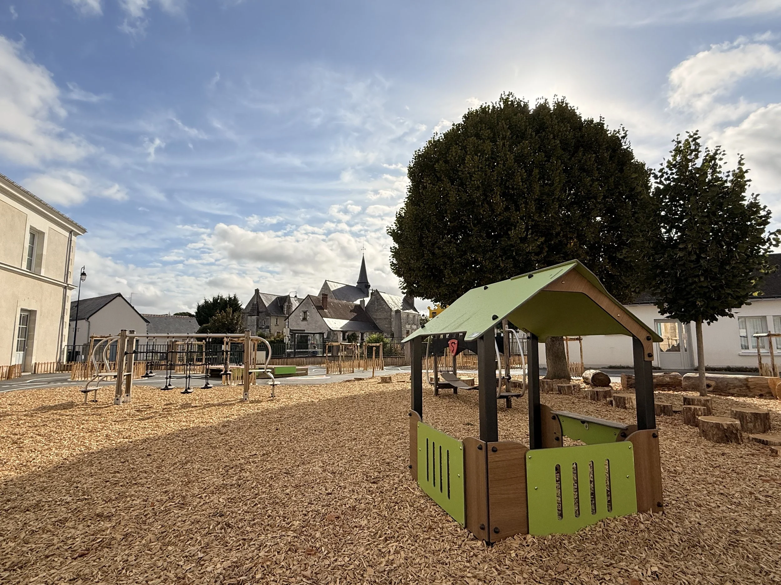 Une aire de jeux pour enfants avec des structures en bois et en métal, entourée de copeaux de bois. Il y a un arbre de grande taille à droite, et des bâtiments résidentiels en arrière-plan. Le ciel est partiellement nuageux.
