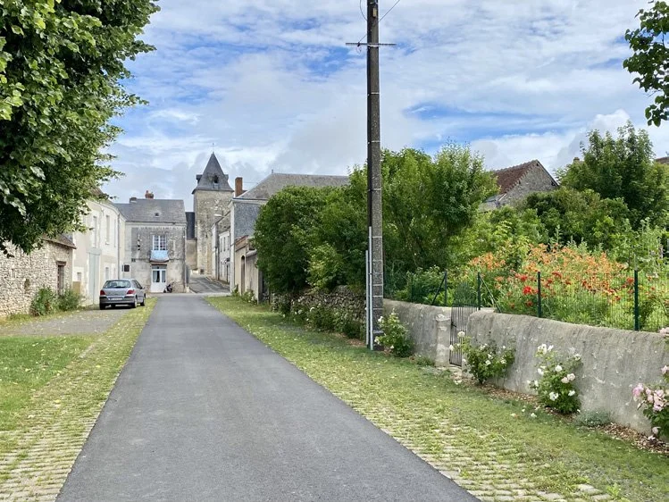 Vue d'une rue rurale avec une route asphaltée, des maisons anciennes, des arbres verts et un ciel partiellement nuageux. En arrière-plan, une église ou un bâtiment historique avec une tour.