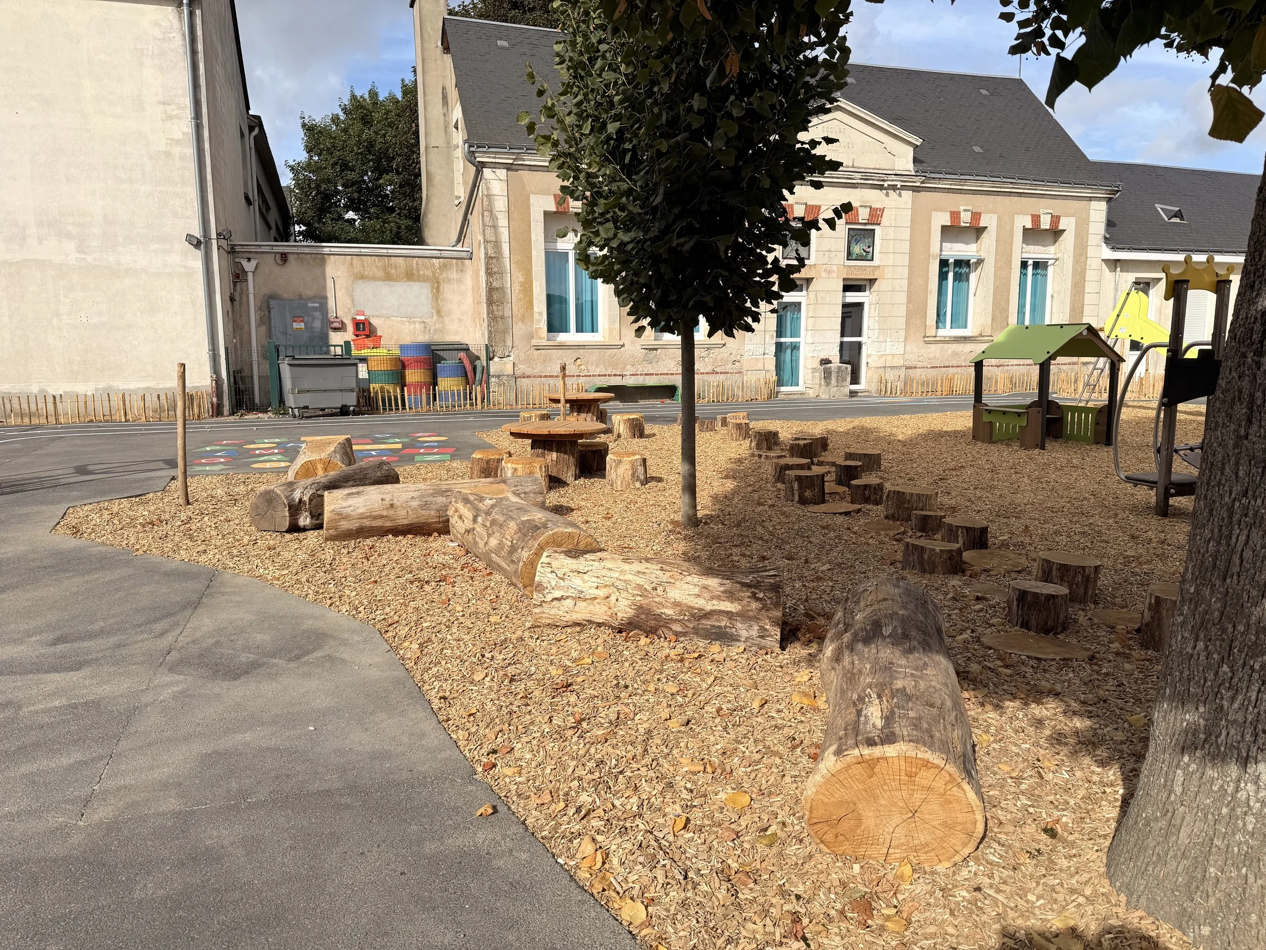 Une cour de récréation pour enfants avec des jeux, des troncs d'arbres disposés comme des sièges, un arbre au centre, au fond un bâtiment en pierre avec des fenêtres et un jardin clôturé.