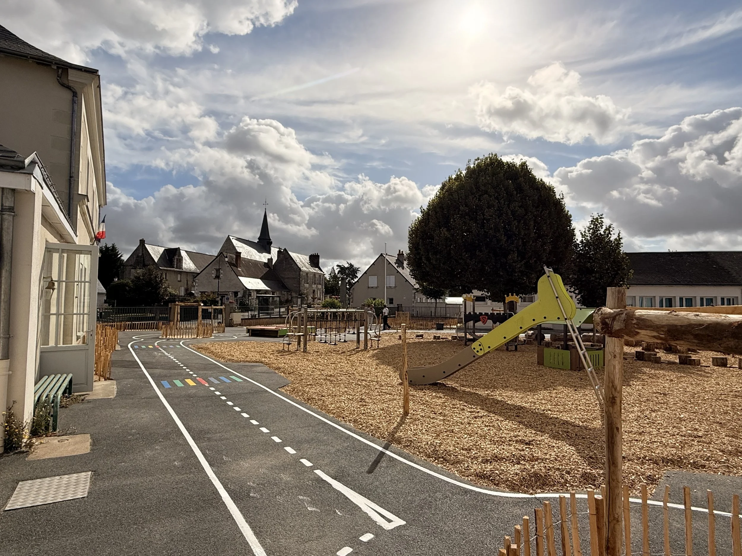 Une cour d'école avec un chemin peint en forme de route, des structures de jeux pour enfants, des bâtiments résidentiels, des arbres et un ciel partiellement nuageux.