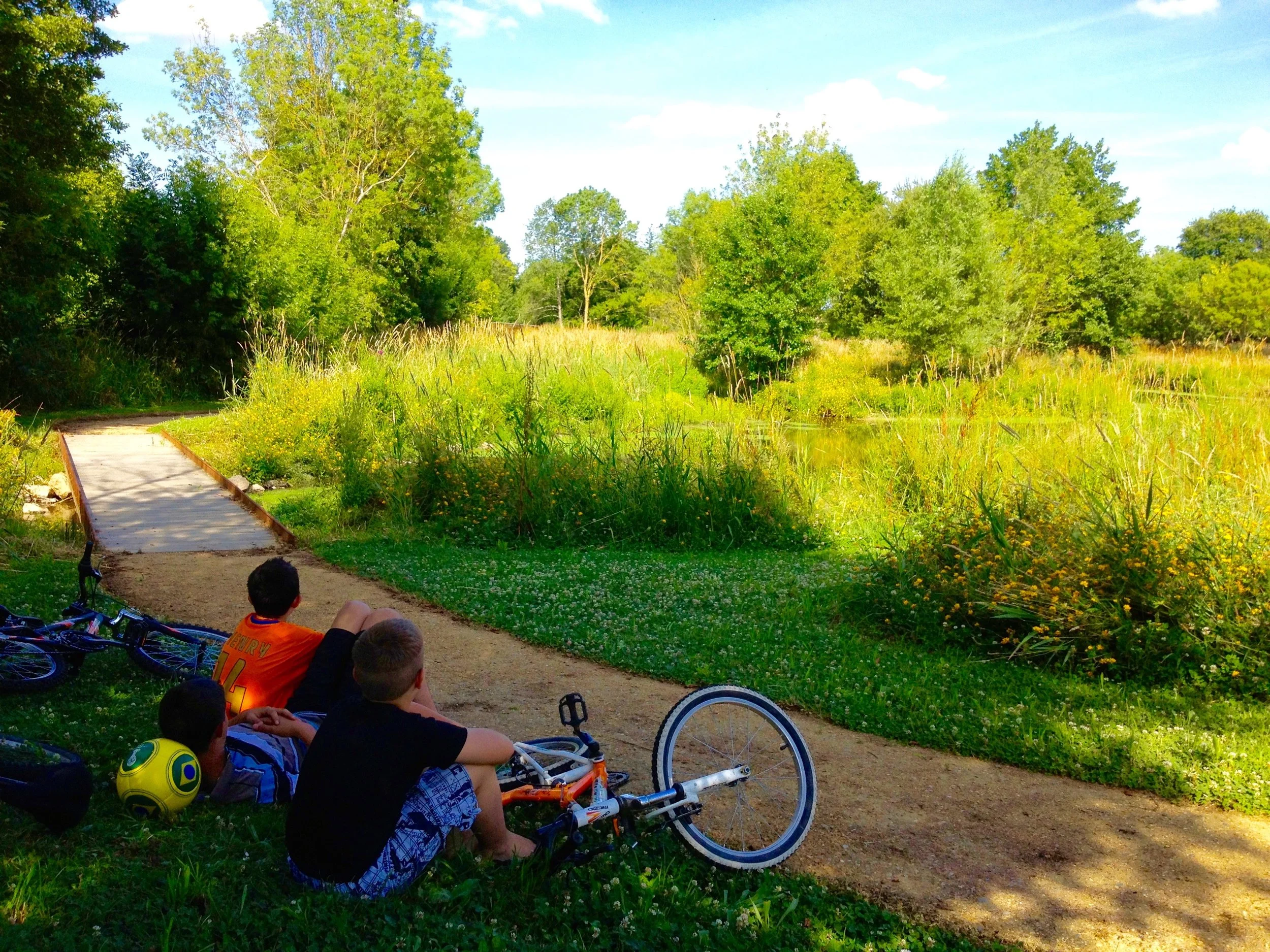 Jeunes enfants assis sur le sol à côté de vélos, certains portant des casques, dans un parc avec une balade en bois et un environnement verdoyant entouré d'arbres. Ils regardent vers un étang ou une rivière au loin.