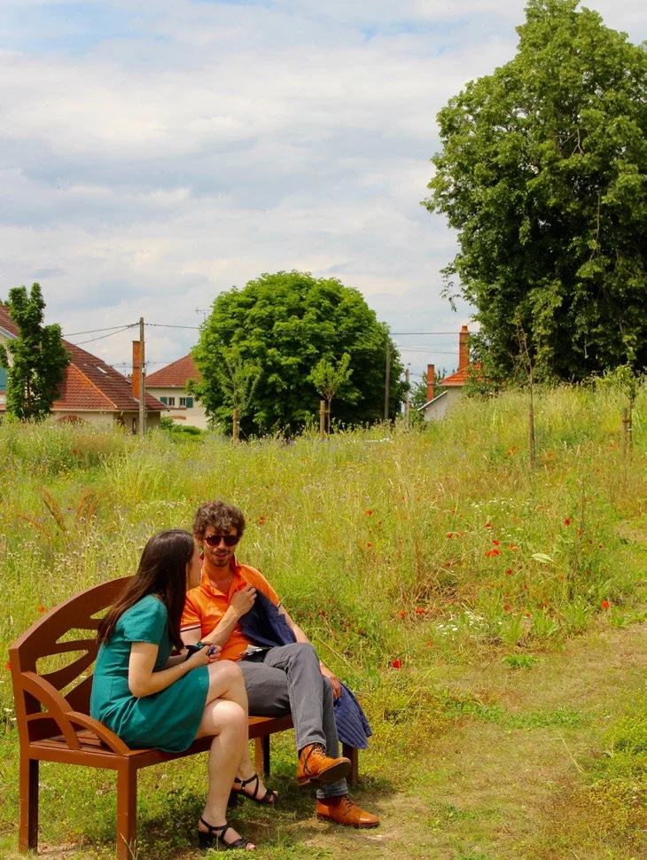 Un homme et une femme assis sur un banc en bois dans un champ d'herbe et de fleurs sauvages, avec des arbres et des maisons en arrière-plan, un ciel partiellement nuageux.