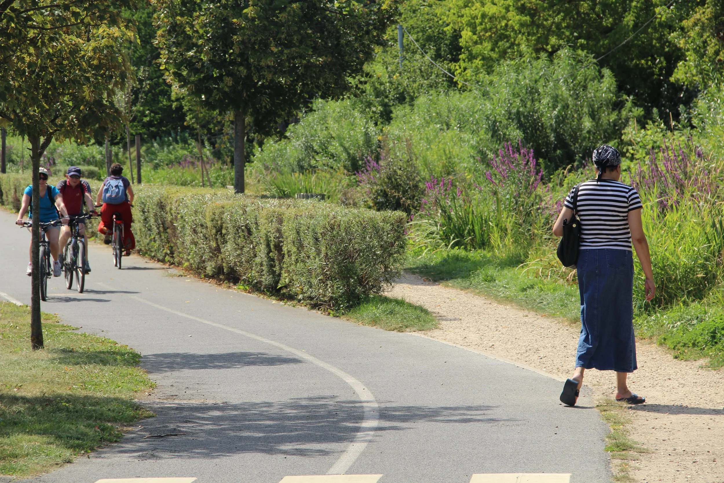 Une femme marche le long d'un sentier pavé, en portant un sac noir, tandis que trois personnes à vélo, portant des casques et des sacs à dos, circulent sur le même chemin. Il y a des arbres et des buissons verts surroundants dans un environnement nat