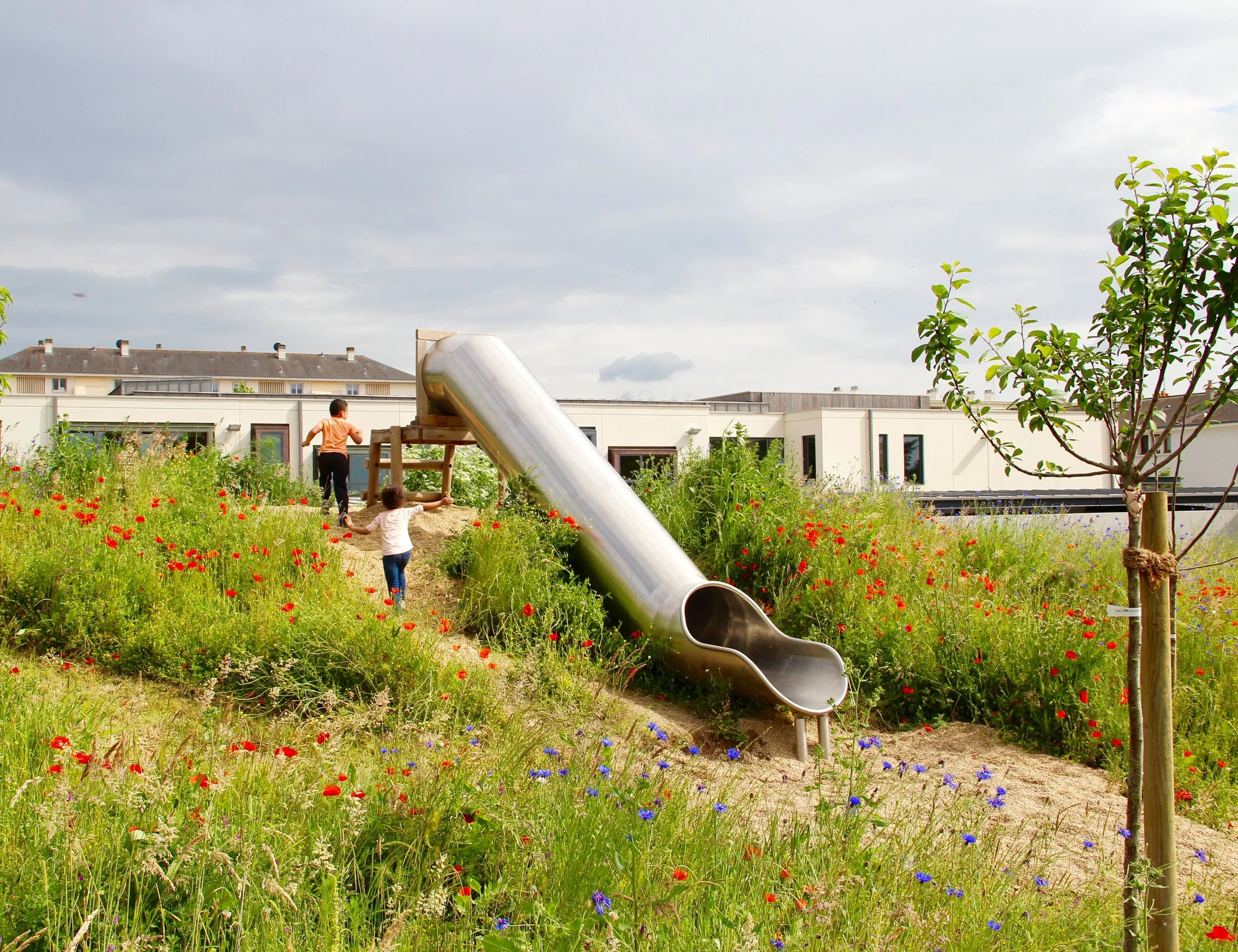 Un jardin avec des fleurs colorées, un enfant jouant près d'une grande glissière en métal, et des maisons modernes en arrière-plan sous un ciel nuageux.