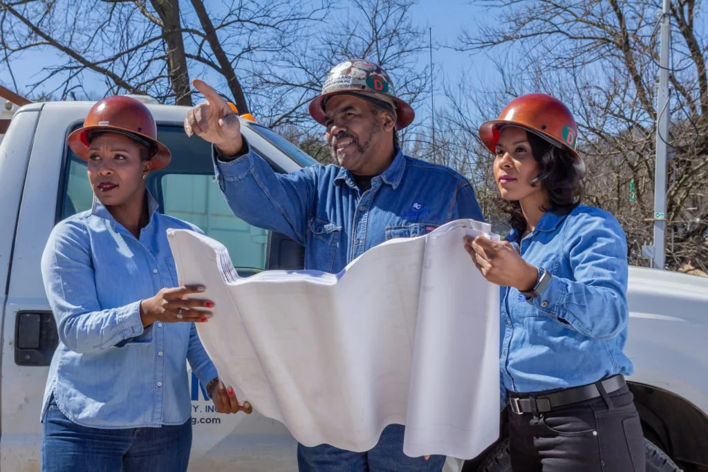 Three utility workers, two women and one man, wearing hard hats and denim shirts, review a large blueprint outdoors near a utility truck with leafless trees in the background.