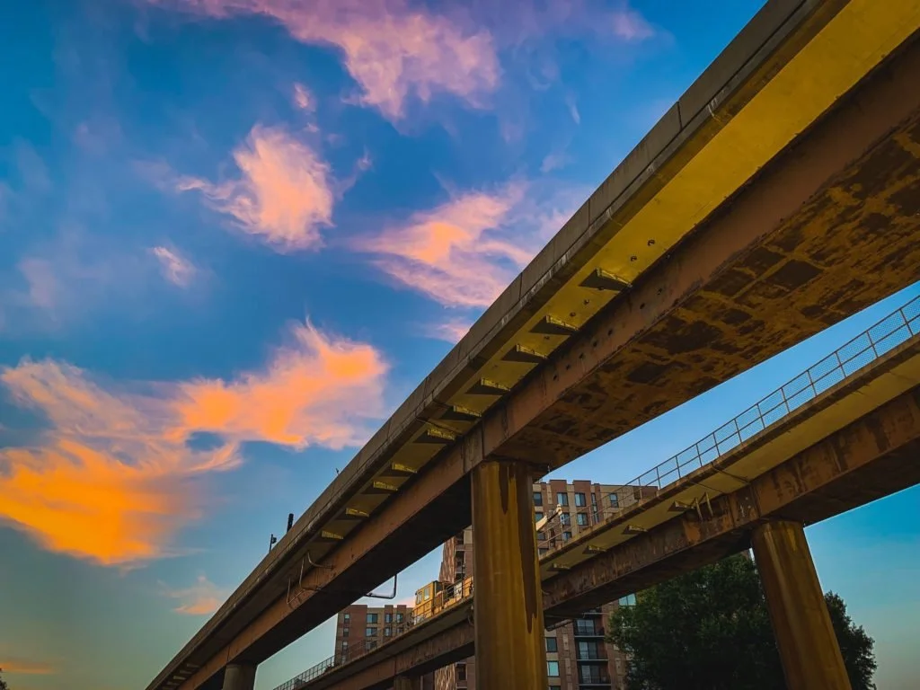 View of an elevated train track or bridge with a building in the background, painted in warm sunset colors with pink and orange clouds in the sky.