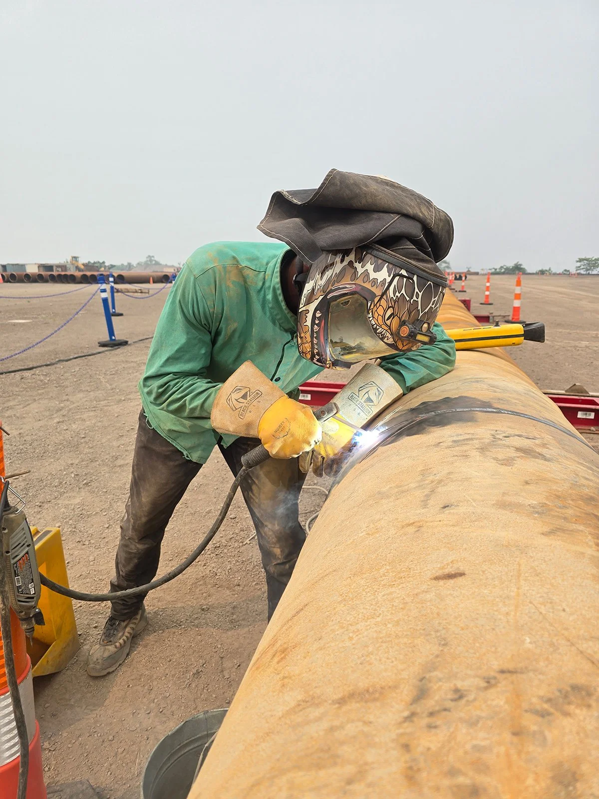Worker welding a large metal pipe outdoors, wearing a welding helmet and gloves, with safety cones and pipes in the background.