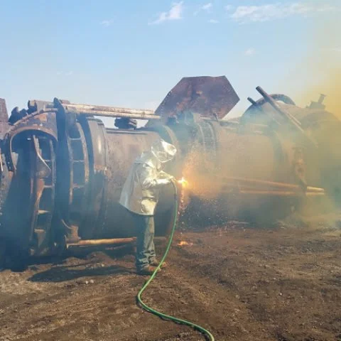 Worker welding on a large, overturned industrial vehicle or machinery in an outdoor setting with a clear sky.