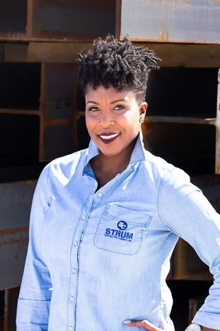 A woman with short curly hair wearing a light blue button-up shirt with a logo, standing outdoors in front of construction materials, smiling at the camera.