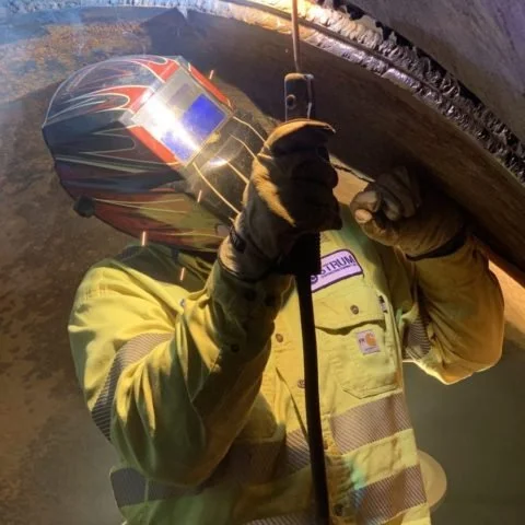 Welding worker wearing a helmet and protective gear welding metal under a bridge or large structure.