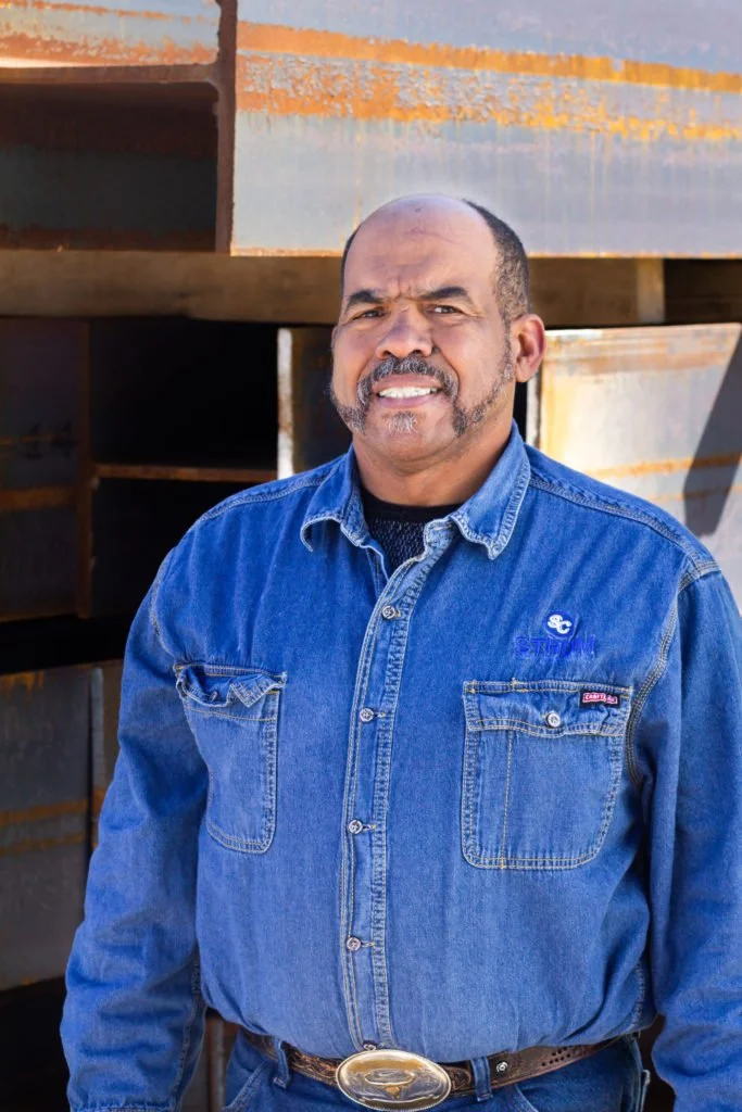 A man with a shaved head and moustache, wearing a blue denim shirt with chest pockets, standing outdoors against a background of rusted metal and wooden structures.