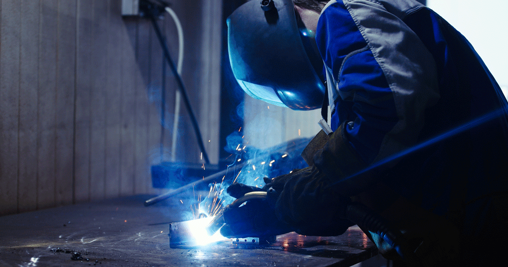 A person welding metal in a workshop, wearing a welding helmet and gloves, sparks flying from the welding process.