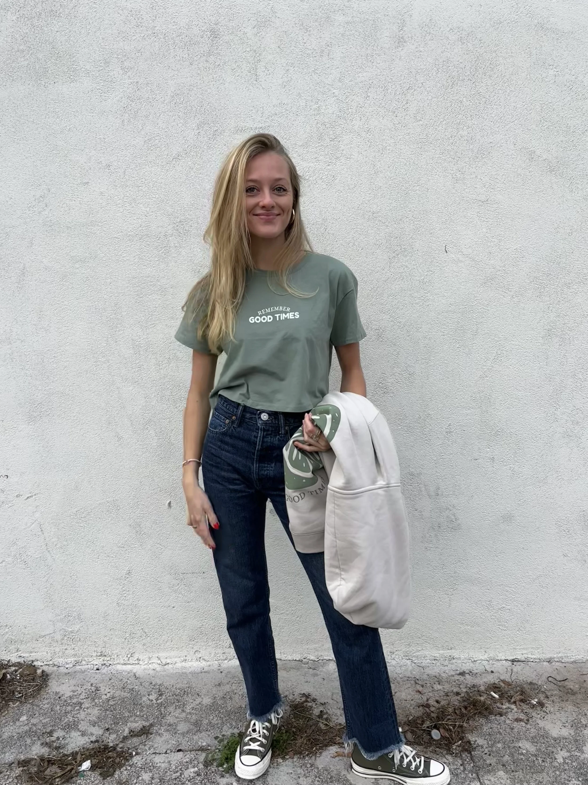 A young woman with long blonde hair smiles while standing against a plain white wall. She is wearing a casual gray T-shirt with the words "Remember Good Times" printed on it, dark jeans, and black and white high-top sneakers. She holds a white hoodie with green design details and a white bag in her left hand. The ground is a mix of concrete and dirt.