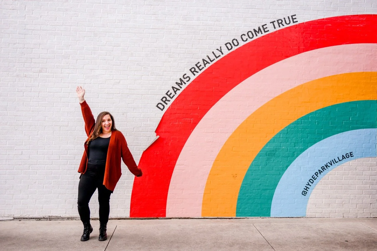 Woman smiling and raising her hand standing next to colorful rainbow mural on brick wall, with the phrase 'Dreams really do come true' written on the red arc.