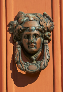 A bronze sculpture of a woman's face with an elaborate hairstyle and decorative necklace, mounted on a wooden background.