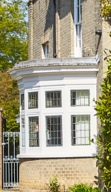 A historic building with a rounded bay window on the ground floor, featuring multiple small-pane windows and decorative brickwork at the base, surrounded by greenery.
