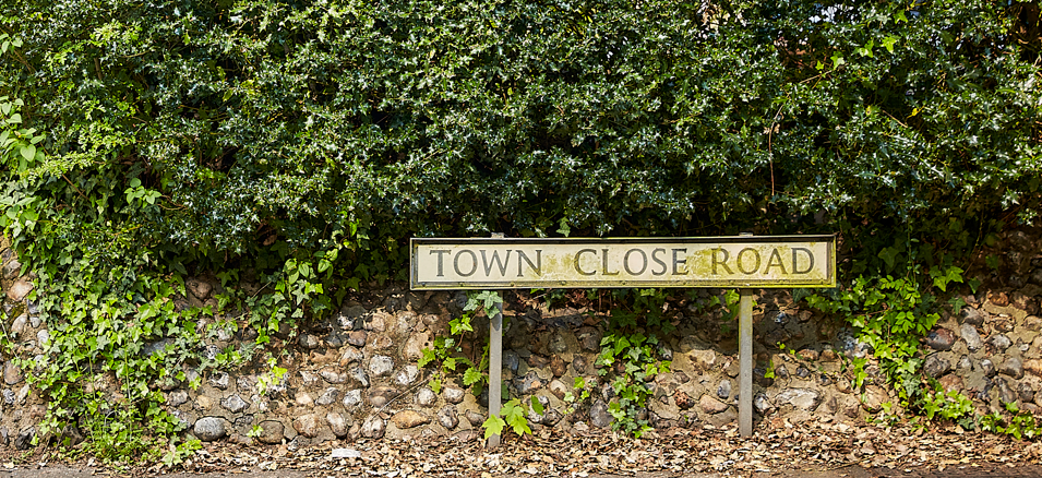 A street sign that reads "Town Close Road" in front of a stone wall and green leafy bushes.