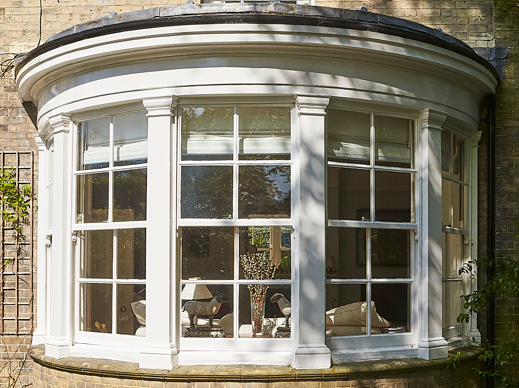 A large, curved bay window with white frames on a brick building, showing a living room inside with chairs, a table, and plants.