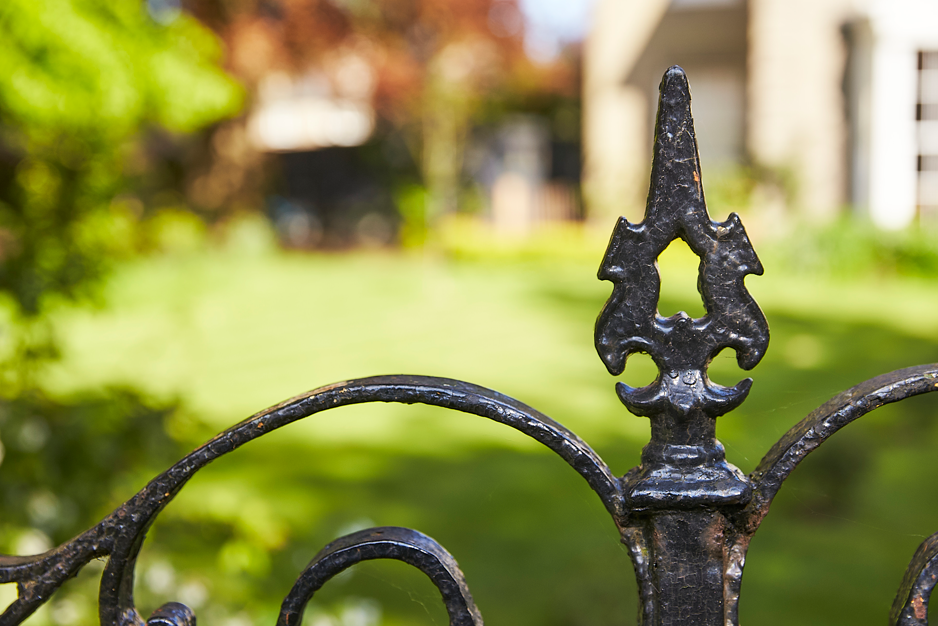 Close-up of a decorative black metal fence with a pointed finial, blurred green yard in the background with trees and a house.