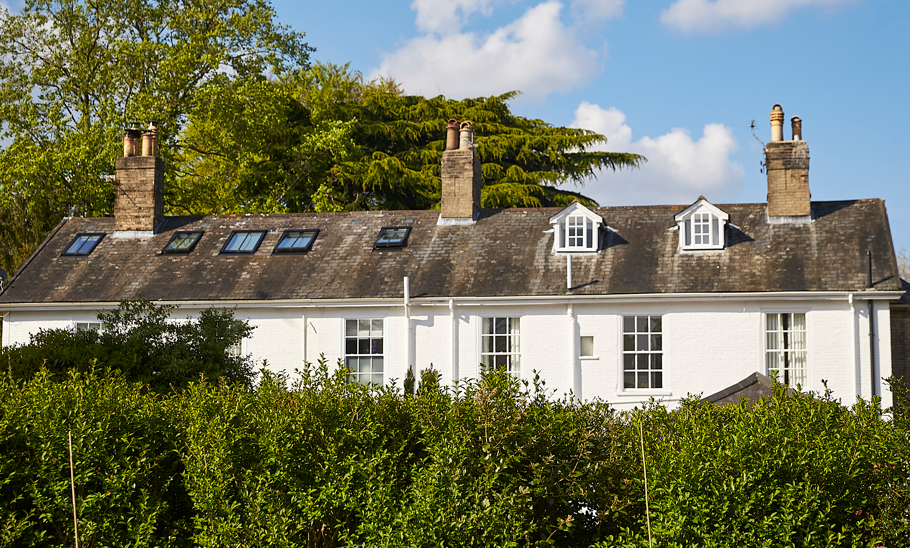 A white house with multiple chimneys, windows, and dormer windows on the roof, surrounded by green bushes and trees under a partly cloudy sky.