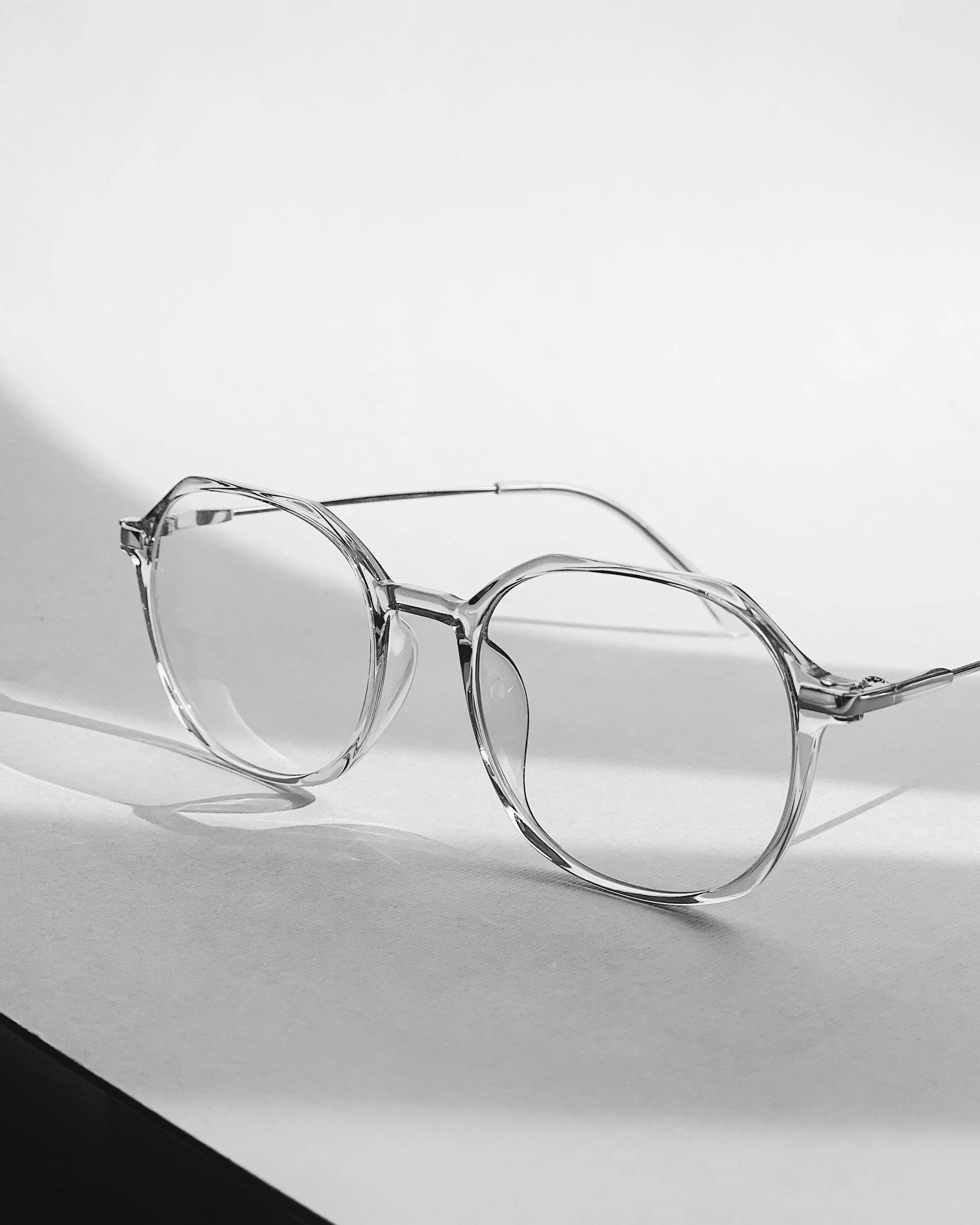 A pair of transparent eyeglasses with round lenses resting on a two-tone background, with a darker bottom edge and a light upper surface.