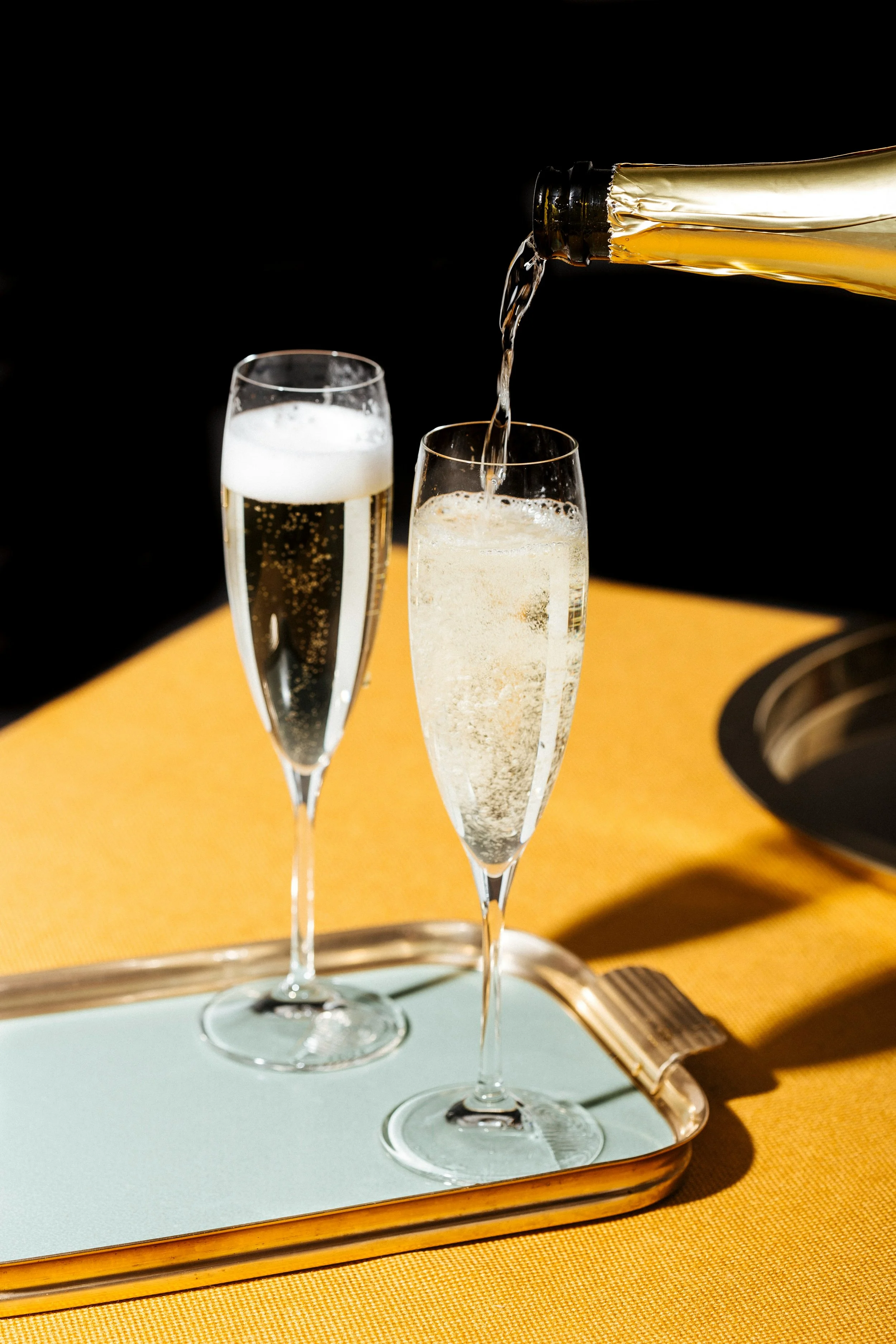 Two champagne flutes on a tray, one being filled with champagne from a bottle against a black background, with a yellow tablecloth underneath.