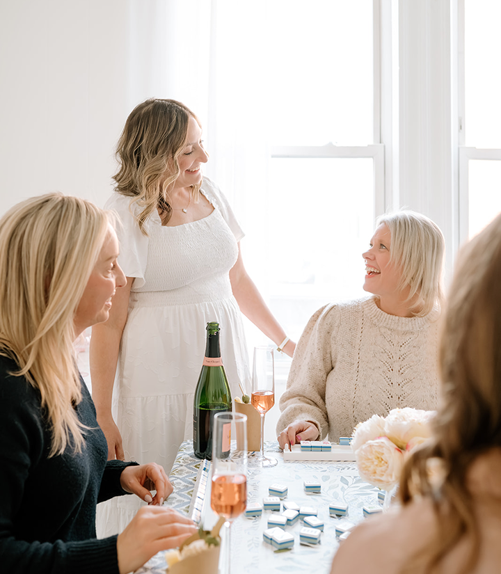 A group of women gathered around a table celebrating, with champagne bottles and glasses, flowers, and dominoes on the table, in a bright room with large windows.