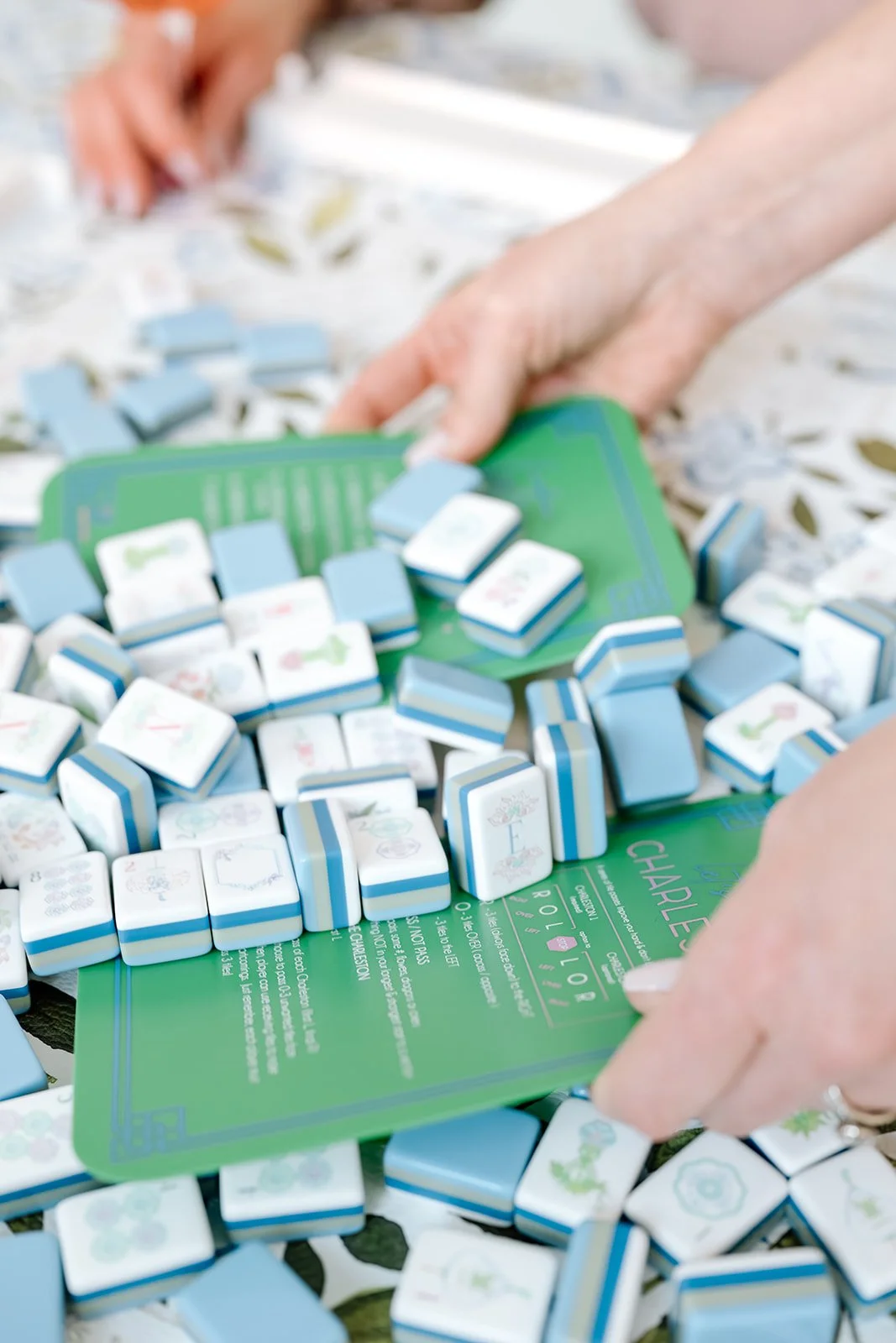 Close-up of hands playing Mahjong with blue and white tiles on a green gameboard.