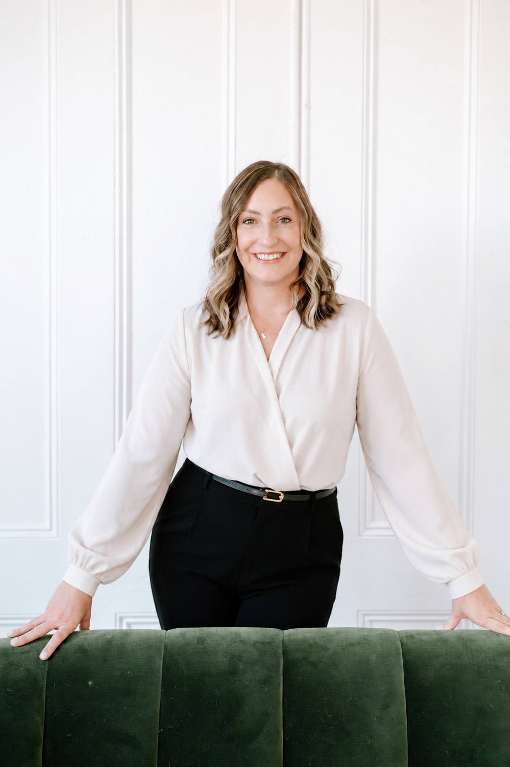 A woman with wavy, shoulder-length brown hair smiling and standing behind a green sofa, dressed in a cream-colored blouse and black pants, against a white paneled wall.