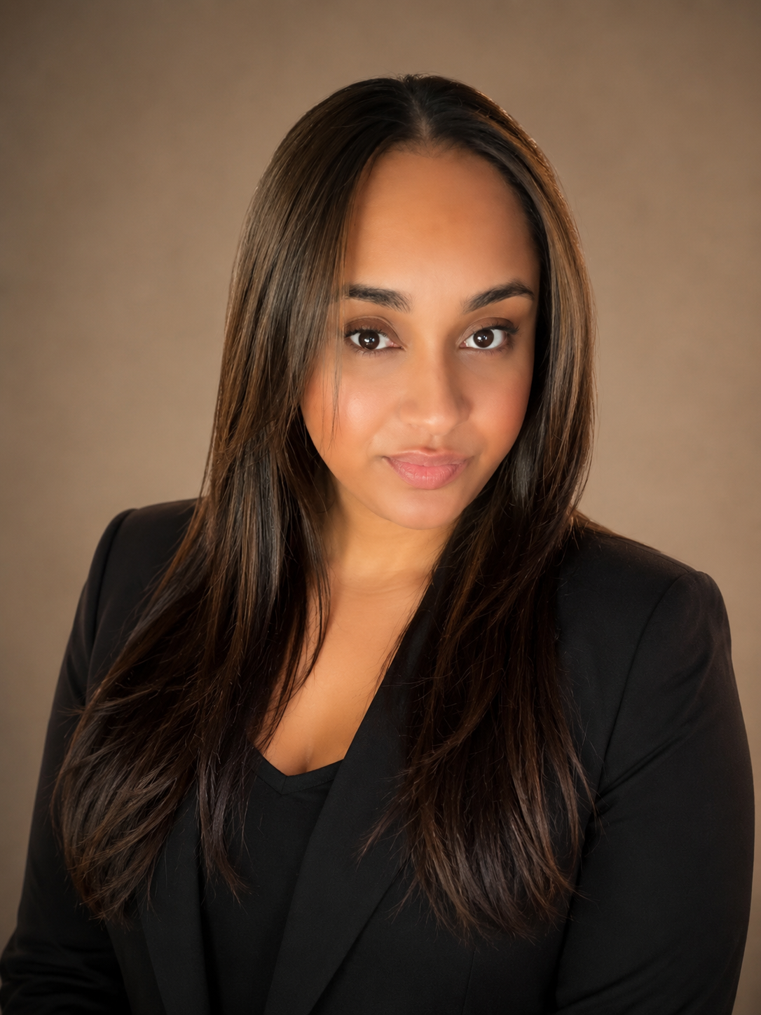 A woman with long brown hair, wearing a black blazer and top, looking confidently at the camera against a neutral background.