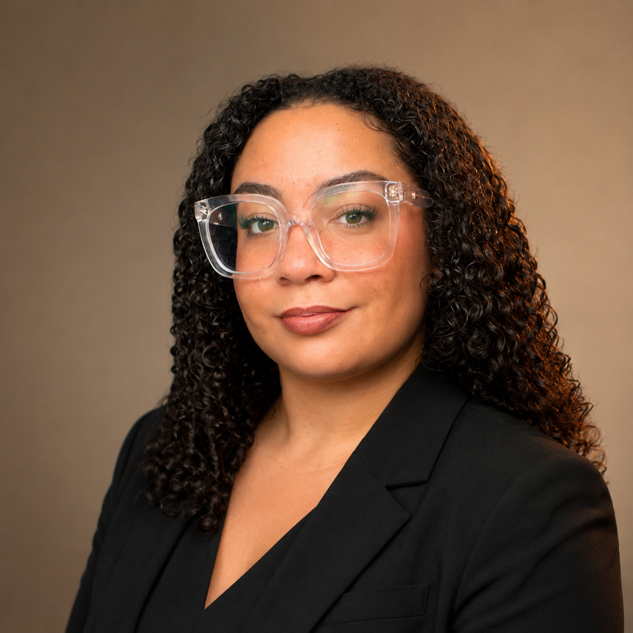 A woman with curly dark hair wearing clear glasses and a black blazer, smiling slightly, in front of a neutral background.