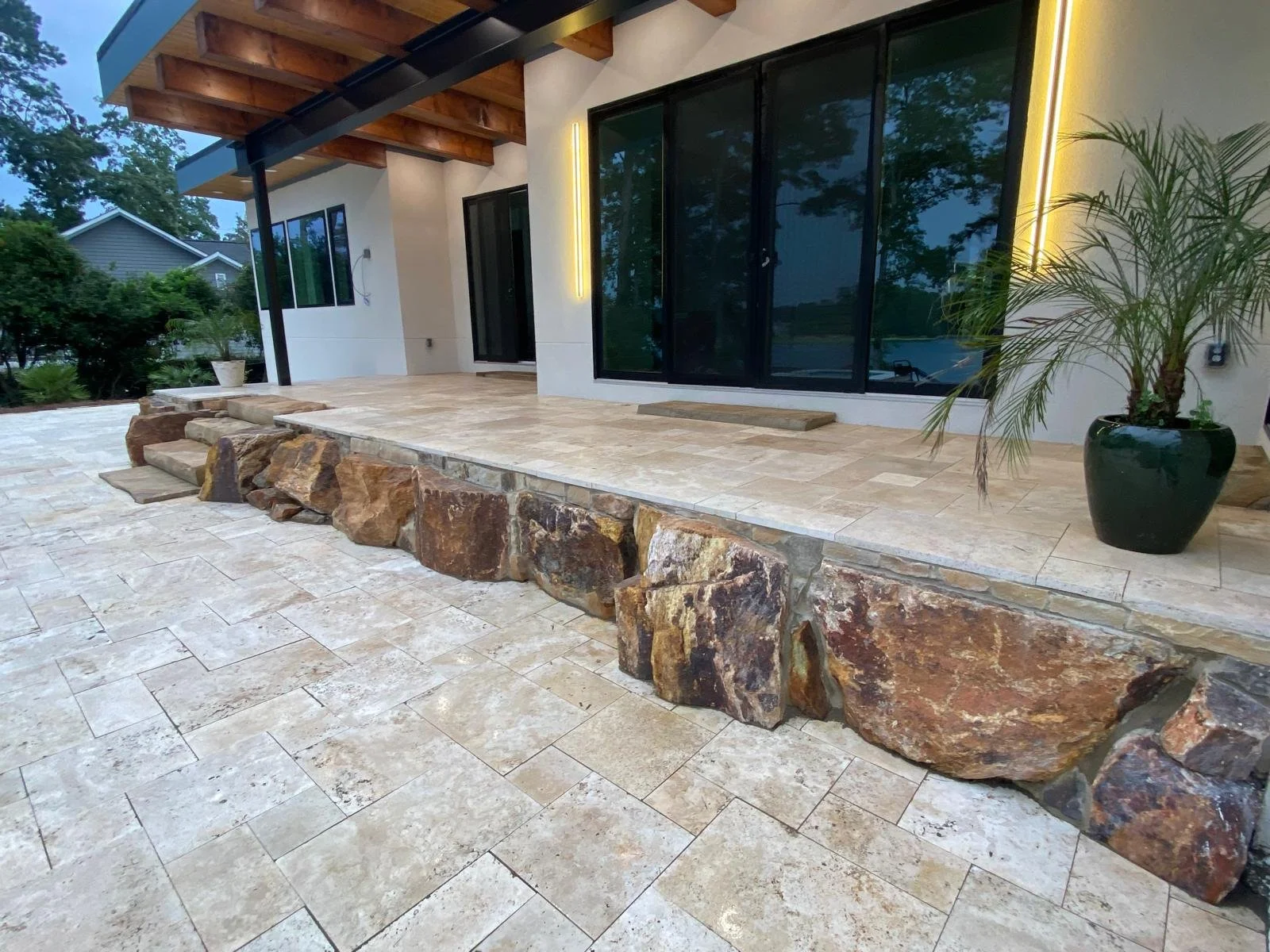 A modern house exterior with a stone patio, large glass sliding doors, a potted plant, and a stone ledge made of large rocks. The patio has a combination of beige and tan tiles, and the house features a wooden overhang.